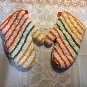 May include: A pair of white oven mitts featuring a textured terry cloth surface with diagonal stripes in orange, yellow, and green. Each mitt includes an orange fabric trim along the edges and a small hanging loop at the top corner.