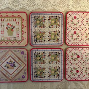 May include: A set of six square coasters featuring various quilt-inspired patterns. The designs include floral baskets, geometric patchwork blocks, and repeating wreath motifs in shades of red, green, yellow, and purple on a light background.