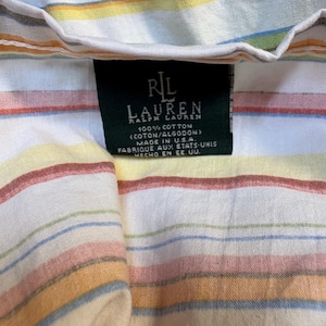 May include: A white cotton shirt featuring horizontal stripes in shades of orange, yellow, and light blue. A black rectangular tag is sewn into the collar area, displaying the text RL LAUREN RALPH LAUREN, 100% COTTON, and MADE IN U.S.A.