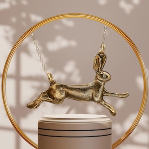 May include: A gold-colored pendant necklace featuring a running hare design. The pendant is suspended from a delicate chain and hangs within a gold circular frame. The hare is detailed with textured fur and a small gem. The necklace rests on a white pedestal.