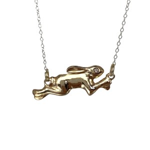May include: A gold-colored pendant necklace featuring a running hare. The hare has a small, clear gemstone eye and is suspended from a delicate silver chain. The pendant is highly detailed and has a polished finish.