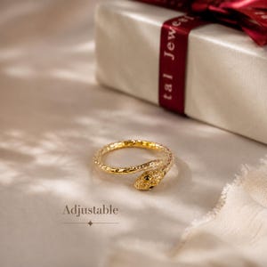 May include: An adjustable gold-toned ring shaped like a coiled snake with a textured surface and dark eyes. The word Adjustable is printed in black text below the ring, which is positioned in front of a white gift box with a red ribbon.