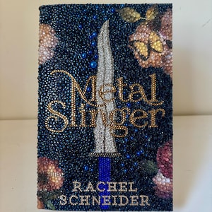 Könnte beinhalten: Ein Buchcover mit dem Titel Metal Slinger von Rachel Schneider, verziert mit dunkelblauen, goldenen und silbernen Strasssteinen. Das Design zeigt einen silbernen Dolch in der Mitte, goldene Schriftzüge und florale Muster mit rosa und goldenen Schmetterlingen.