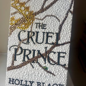 Könnte beinhalten: Ein Buchcover für The Cruel Prince von Holly Black mit einem weißen, texturierten Hintergrund aus kleinen Perlen. Das Design zeigt eine goldene Perlenkrone, dunkelbraune Perlenzweige und eine schwarze Perlenbeschriftung für den Titel.