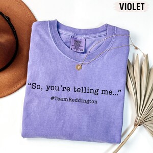 以下が含まれることがあります： 「So, you're telling me...」および「#TeamReddington」という黒い文字がプリントされた、バイオレットカラーの半袖クルーネックTシャツ。襟の内側にはComfort Colorsのブランドラベルがあり、ゴールドトーンのネックレスが添えられています。