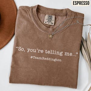 以下が含まれることがあります： エスプレッソカラーの半袖Tシャツで、クルーネック仕様です。シャツには「So, you're telling me...」という白い文字がプリントされており、その下に「#TeamReddington」と記されています。生地の上にはシルバー調のチェーンネックレスが置かれています。