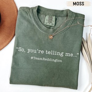 以下が含まれることがあります： モスグリーンの半袖クルーネックTシャツ。シャツには「So, you're telling me...」と「#TeamReddington」という白い文字がプリントされています。小さな円形のペンダントが付いた細いゴールドトーンのチェーンネックレスがTシャツの上に置かれています。