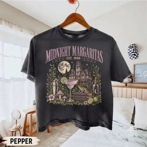 May include: A charcoal gray t-shirt featuring a graphic print with the text "MIDNIGHT MARGARITAS EST. 1998". The design includes a large house, a full moon, a cocktail glass, a shaker, a frog on books, and foliage in shades of green and pink.