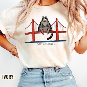 May include: An ivory t-shirt featuring a graphic illustration of a grey tabby cat sitting on the red Golden Gate Bridge. Below the bridge, the text SAN FRANCISCO is printed in black. The cat has wide eyes and a striped pattern on its body.