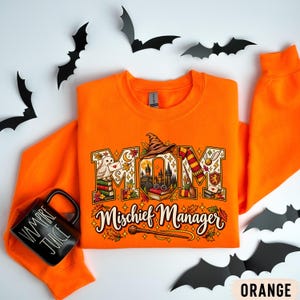 May include: An orange long-sleeve sweatshirt featuring a graphic print of the word MOM with wizarding-themed illustrations inside the letters, including an owl, books, and a castle. Below, the text reads Mischief Manager with a wand graphic.
