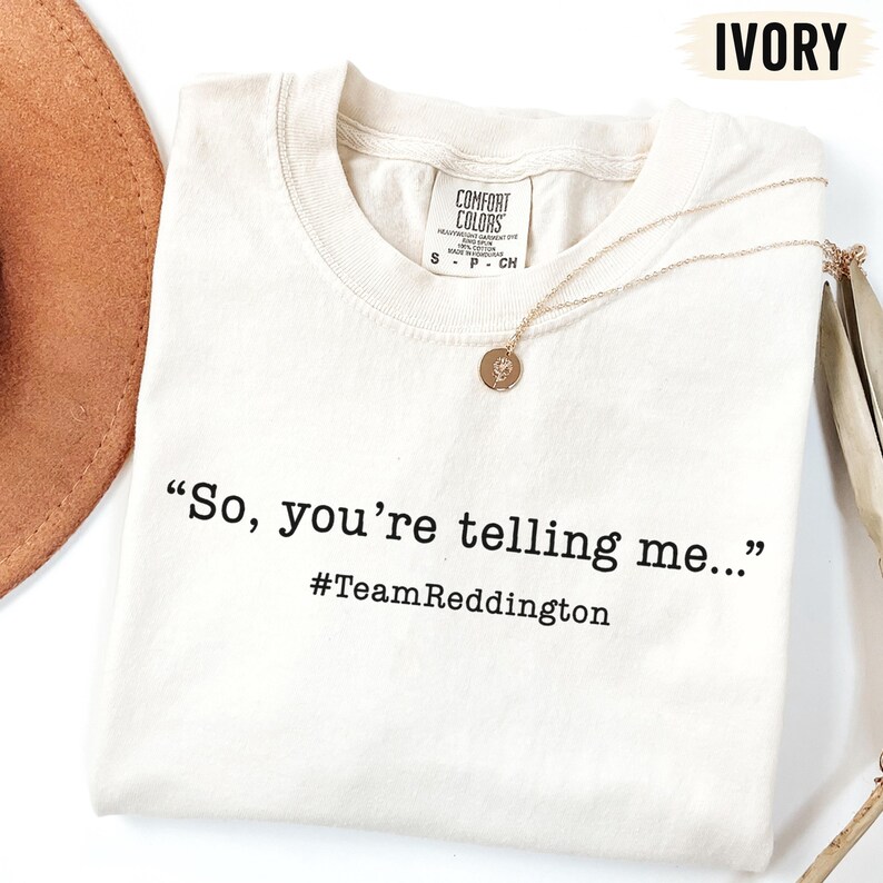 以下が含まれることがあります： アイボリーの半袖Tシャツで、黒い文字で「So, you're telling me...」と「#TeamReddingtion」がプリントされています。Tシャツの内側の襟元にはComfort Colorsのブランドラベルがあり、繊細なゴールドトーンのネックレスが添えられています。