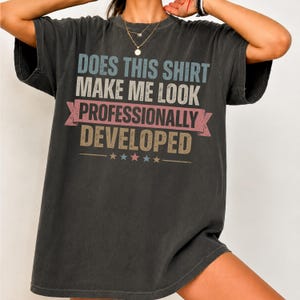 Peut inclure: Un t-shirt gris anthracite à manches courtes avec le texte "DOES THIS SHIRT MAKE ME LOOK PROFESSIONALLY DEVELOPED" dans une police vieillie et multicolore. Le motif comprend une bannière rose et cinq petites étoiles en bas.
