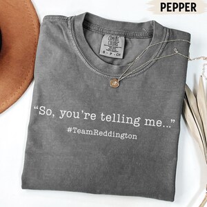 以下が含まれることがあります： クルーネックのチャコールグレーの半袖Tシャツで、Comfort Colorsのブランドラベルが付いています。シャツには白い文字で「So, you're telling me...」とプリントされており、その下に白いフォントで「#TeamReddington」というハッシュタグが記されています。