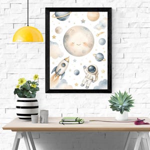 Könnte beinhalten: Ein gerahmter Kunstdruck mit einer Aquarellillustration eines lächelnden Mondes, einer weißen Rakete mit orangefarbenem Abgasstrahl und einem im Weltraum schwebenden Astronauten, ergänzt durch verschiedene Planeten, Sterne und weiche Wolken.