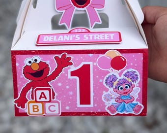 May include: A white cardboard favor box featuring a pink panel with a large red number one, Elmo, Abby Cadabby, and ABC blocks. A pink bow with Elmo's face sits on top, and a sign reading DELANI'S STREET is attached to the front of the box.