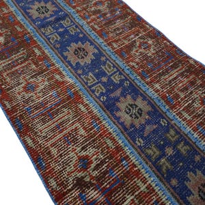 May include: A rectangular runner rug featuring a central deep blue band decorated with repeating geometric star motifs in beige and brown. The side panels display a distressed pattern of red, brown, and cream tones with intricate linear details.
