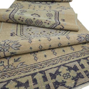 May include: A long, rectangular runner rug featuring a beige base color with intricate patterns in dark charcoal and muted sage green. The design includes geometric borders and central floral motifs, with the rug folded over itself in sections.