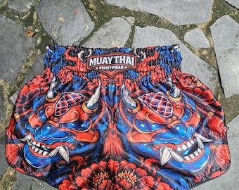 Pode incluir: Calções de combate Muay Thai com uma estampa gráfica vibrante de duas máscaras oni vermelhas e azuis com presas brancas afiadas. O cós elástico exibe um emblema preto com o texto branco MUAY THAI FIGHTWEAR.