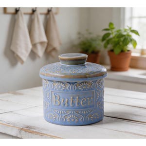 Peut inclure: Un pot à beurre cylindrique en céramique avec couvercle, doté d'un vernis bleu clair et d'un motif floral en relief répété. Le mot Butter est centré sur le devant en lettres saillantes. Le bord et le couvercle ont une finition brun doré.