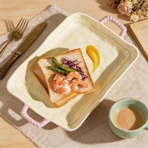 Può includere: Un vassoio da portata rettangolare in ceramica color crema con due manici intrecciati rosa chiaro. Il vassoio contiene due fette di pane tostato guarnite con gamberi cotti, punte di asparagi verdi e cavolo viola, accompagnate da una salsa gialla.