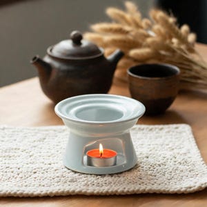 Peut inclure: Un brûle-parfum en céramique blanche avec un bassin supérieur circulaire et une base creuse contenant une bougie chauffe-plat allumée avec une flamme orange visible. Le brûleur présente une finition lisse et brillante et une base large et stable.