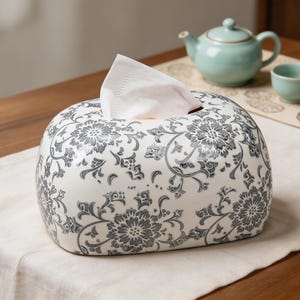 May include: A rectangular ceramic tissue box cover featuring a white base with a dark gray floral pattern. The design consists of intricate, repeating flower motifs and swirling vine details across the entire exterior surface of the container.