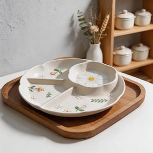 Può includere: Un piatto da portata diviso in ceramica color crema con bordo smerlato e una sezione centrale a forma di ciotola. Il piatto presenta motivi floreali dipinti a mano, tra cui piccole margherite, fiori arancioni e foglie verdi.