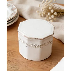 May include: A white ceramic canister with an octagonal shape and a lid. The canister features a raised floral relief pattern around the middle and distressed brown edges along the rim of the lid and the base, providing a textured appearance.