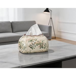 May include: A cream-colored ceramic tissue box cover with a rounded, organic shape. The surface features a delicate floral pattern in shades of green and soft pink. A single white tissue is pulled through the top opening of the container.