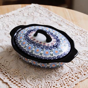 May include: An oval ceramic casserole dish with a matching lid, featuring a black rim and handles. The surface is decorated with a repeating pattern of light blue and orange daisy-like flowers, accented by dark blue swirling scroll motifs.