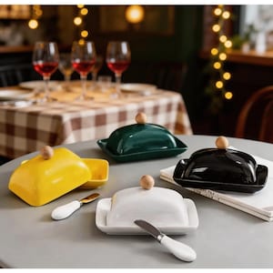 May include: Four rectangular ceramic butter dishes with lids in yellow, dark green, black, and white. Each lid features a small, round, light-colored wooden knob handle. Two matching butter knives with white handles are placed on the table surface.