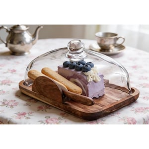Puede incluir: Una bandeja de madera rectangular con una cúpula de cristal transparente. Dentro, una porción de pastel morado con arándanos frescos y crema blanca se encuentra junto a dos bizcochos y un pequeño utensilio de madera para servir.