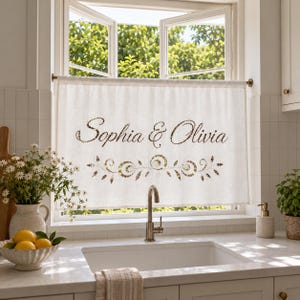 May include: A white cafe curtain hanging on a rod over a kitchen sink, featuring the text "Sophia & Olivia" in a brown script font, centred above a decorative brown floral vine pattern printed on the fabric surface of the window treatment.