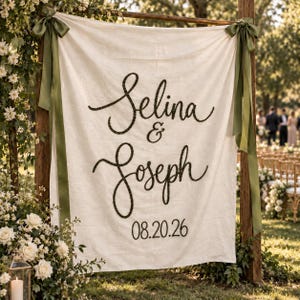 May include: A cream-coloured fabric wedding sign featuring the black script names Selina & Joseph and the date 08.20.26. The banner is suspended from a wooden frame with olive green ribbons tied at the top corners, creating a decorative display.