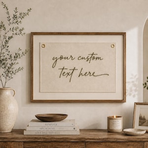 Könnte beinhalten: Ein rechteckiges Wandbild mit einer cremefarbenen Stoffplatte, auf der der dunkelgrün gestickte Text "your custom Text here" zu sehen ist, befestigt mit zwei goldfarbenen Metallösen in einem schlichten Holzrahmen.