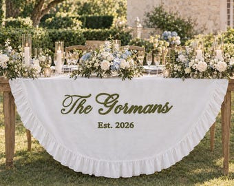 May include: A white semi-circular table skirt with a ruffled edge, featuring the dark green embroidered text The Gormans and Est. 2026. The table is decorated with white and blue floral arrangements, tall glass candles, and clear glassware.
