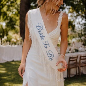 May include: A white sash featuring long fringe trim along the edge and the text Bride To Be embroidered in blue script lettering. The sash is worn diagonally across the torso over a white dress with ruffled shoulder straps.