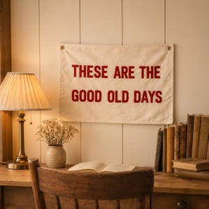 Könnte beinhalten: Ein rechteckiger Wandbehang aus cremefarbenem Stoff mit dem in Rot gestickten Text "THESE ARE THE GOOD OLD DAYS" in einer fetten serifenlosen Schrift. Das Banner verfügt über Metallösen in den oberen Ecken zur Befestigung.