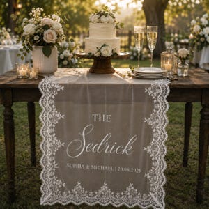 Op de afbeelding: Een doorschijnende witte kanten tafelloper met de tekst THE Sedrick SOPHIA & MICHAEL | 20.06.2026, gedrapeerd over een houten tafel met een witte taart van twee lagen, bloemstukken, twee champagneglazen en een glazen lantaarn met kaars.