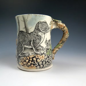 May include: A ceramic mug with a detailed illustration of an animal, possibly a weasel or ferret, in black and white. The handle has a textured, earthy design. The mug's base features a pattern of small, rounded stones.