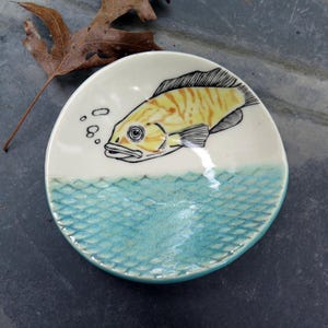 May include: A small, round ceramic dish with a hand-painted fish design. The fish is yellow with black outlines, above a turquoise water pattern. The dish has a white upper section and a turquoise rim.