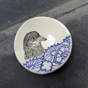 May include: A round, off-white ceramic bowl featuring a detailed black and white illustration of a bird head with a pink eye. The bowl is decorated with a vibrant blue, intricate lace-like pattern that curves along the lower edge of the interior.