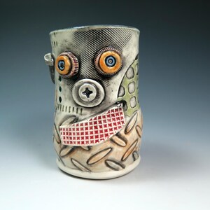 May include: A cylindrical ceramic mug featuring a textured surface with a mechanical aesthetic. The mug displays two orange-rimmed eyes, a central screw-like nose, and a red grid-patterned mouth. The base has a raised diamond plate pattern.