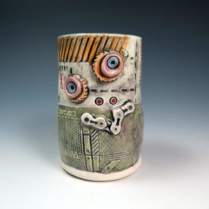 May include: A cylindrical ceramic mug featuring a textured, industrial-inspired surface with a green metallic grid pattern on the lower half. The upper section is off-white with orange stripes, two protruding pink eye-like circles, and a silver chain.