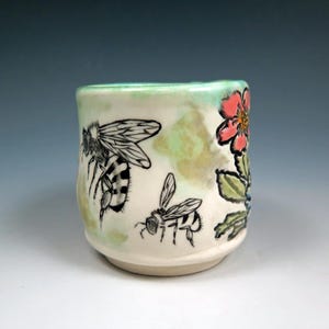 May include: A white ceramic mug with a green glaze and floral and bee designs. The mug features a pink flower, green leaves, and two black and white bees.