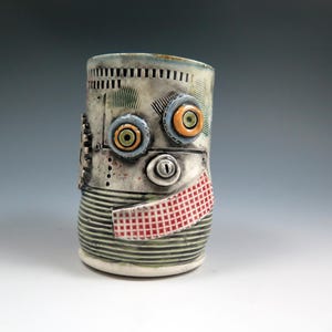 May include: A ceramic mug with a robot face. The mug is decorated with green, blue, red, and orange details. The mug has a textured surface and a striped pattern.