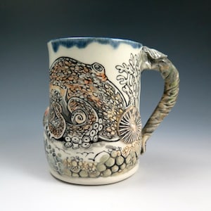 May include: A ceramic mug with a detailed octopus design. The mug features a cream-colored base with a blue rim and a textured handle. The octopus is depicted in shades of brown, orange, and black, with coral and sea life accents. The mug is a unique piece of art.