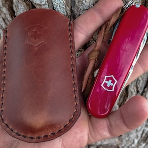 May include: A brown leather pouch with a stamped shield logo and a red multi-tool pocket knife featuring a white shield emblem and a brown leather lanyard. The pouch has visible dark stitching along the edges and a rounded top flap design.