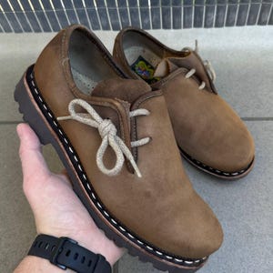 Op de afbeelding: Een paar bruine leren schoenen met een ronde neus, dikke donkerbruine profielzolen met witte contraststiksels langs de rand, en lichtbeige veters gestrikt in een strik. De binnenvoering is licht van kleur met een klein kleurrijk logolabel.