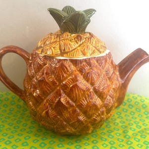May include: A ceramic teapot shaped like a pineapple with a textured brown body and a removable lid featuring a green leafy top. The teapot has a curved handle on the left side and a spout on the right side, with a glossy finish throughout.
